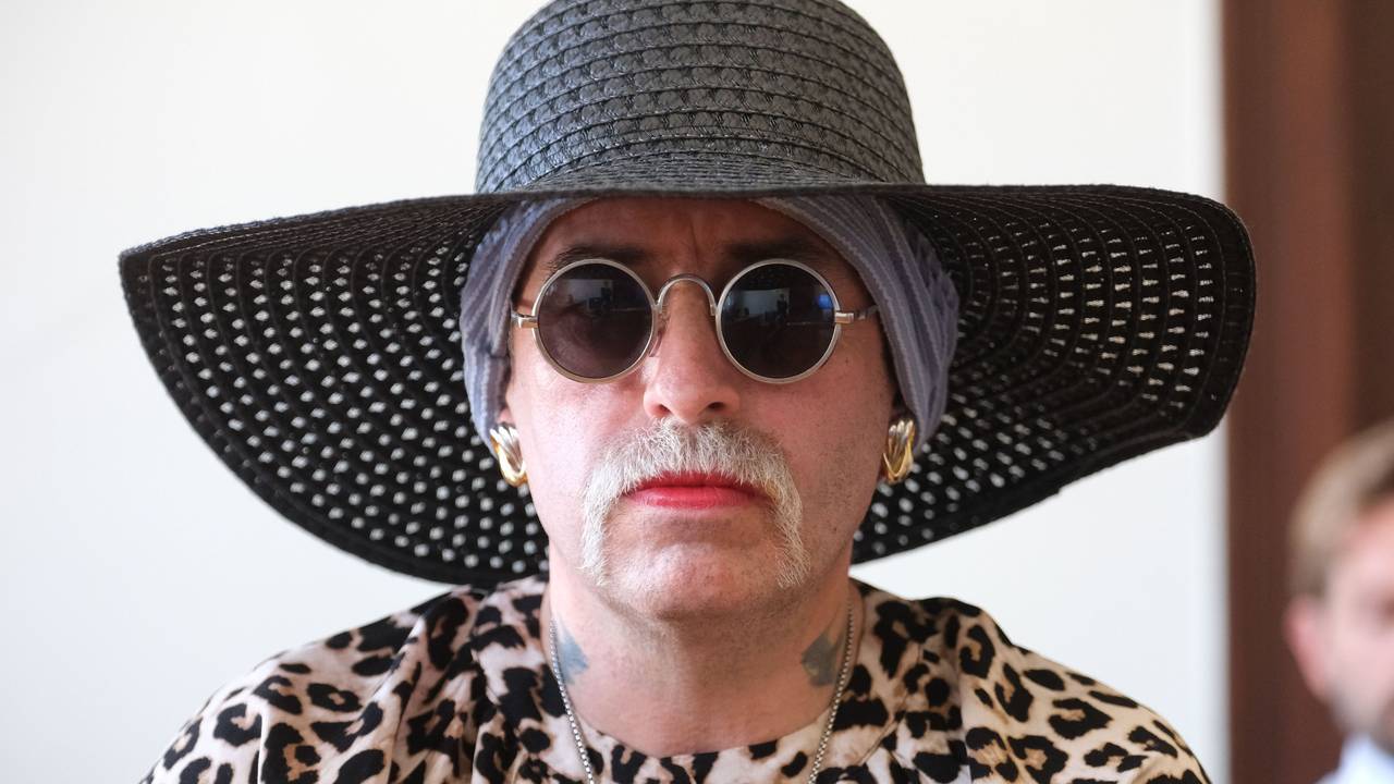 Eine Frau trägt Hut, Brille und Schnurbart, ihre Lippen sind rot geschminkt.