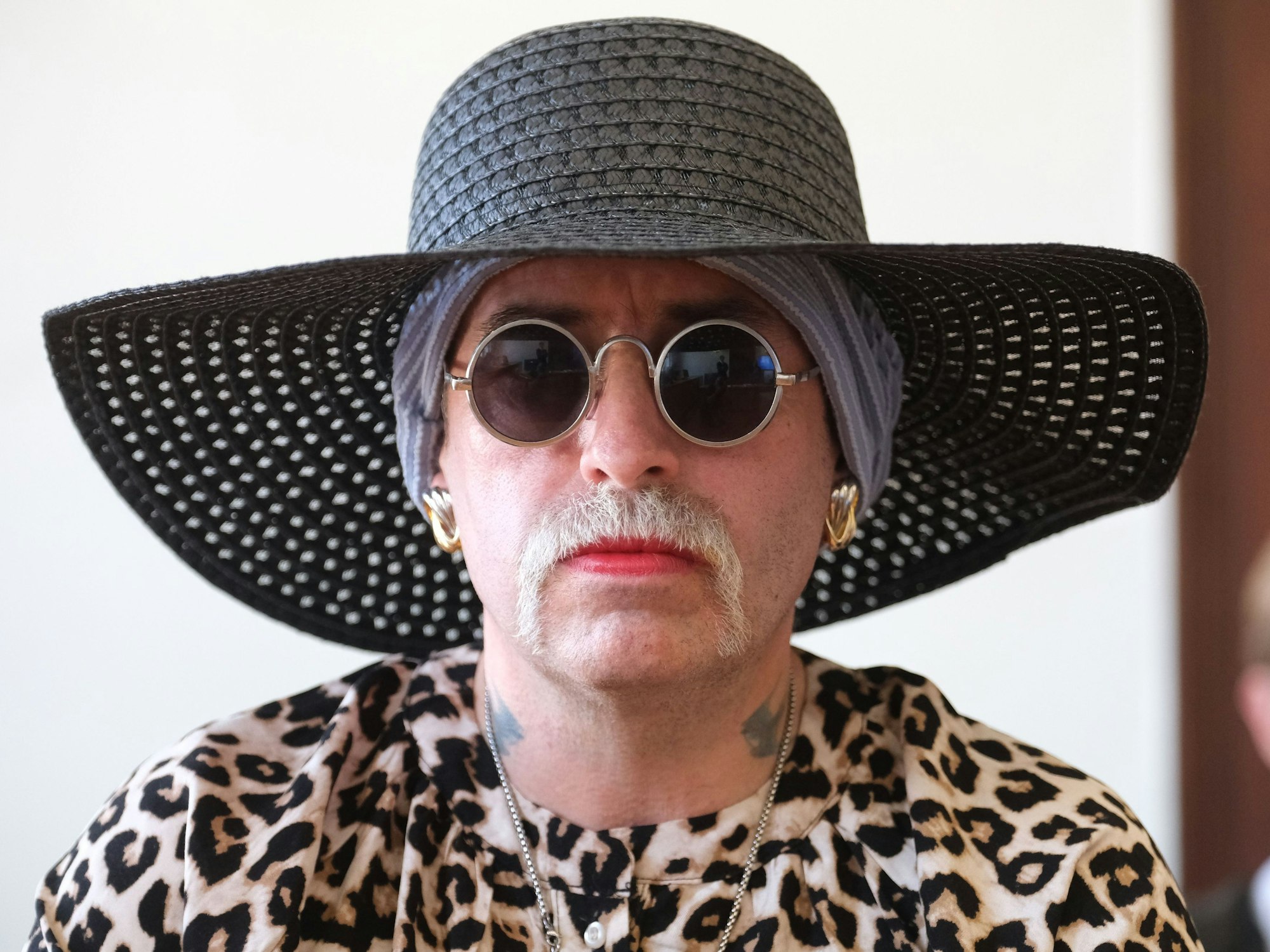 Eine Frau trägt Hut, Brille und Schnurbart, ihre Lippen sind rot geschminkt.