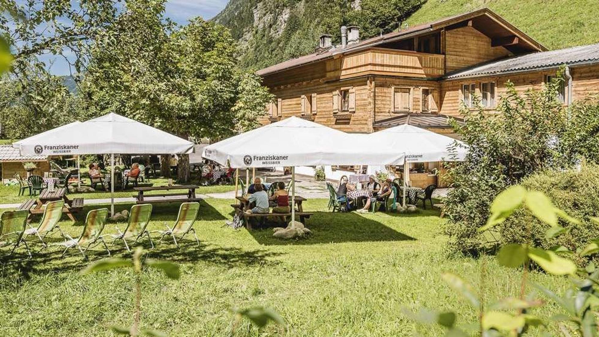 Eine Berghütte mit Biergarten auf einer Wiese inmitten hoher Berge