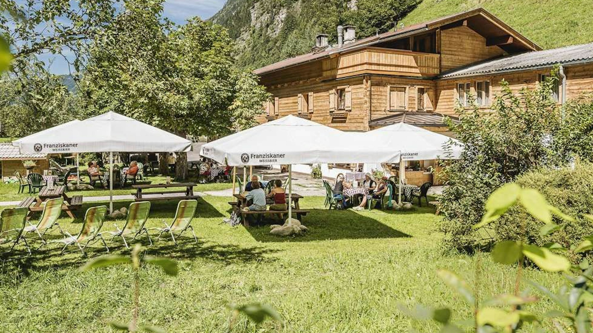 Eine Berghütte mit Biergarten auf einer Wiese inmitten hoher Berge