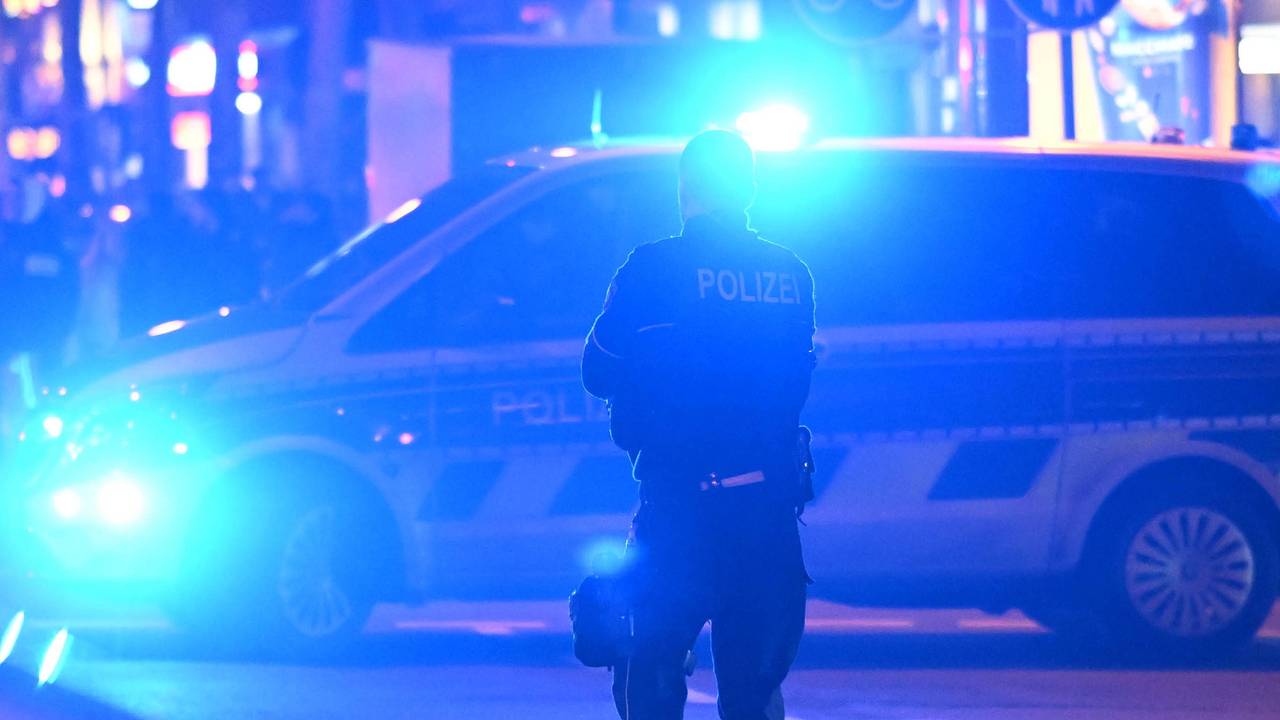 Ein Polizist steht vor einem Polizeifahrzeug, bei dem das Blaulicht eingeschaltet ist.