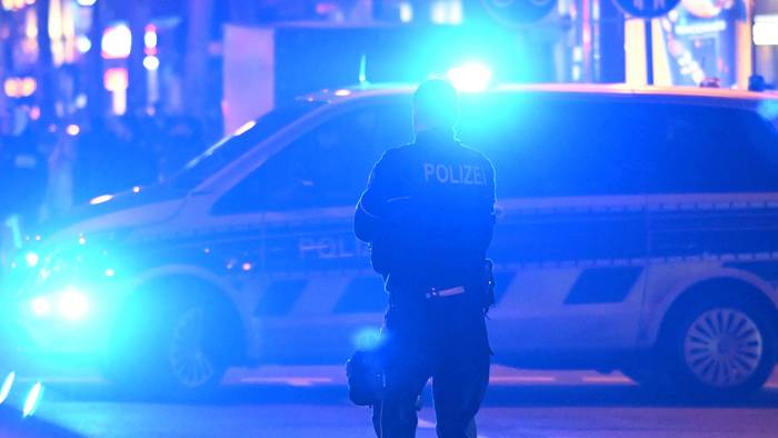 Ein Polizist steht vor einem Polizeifahrzeug, bei dem das Blaulicht eingeschaltet ist.