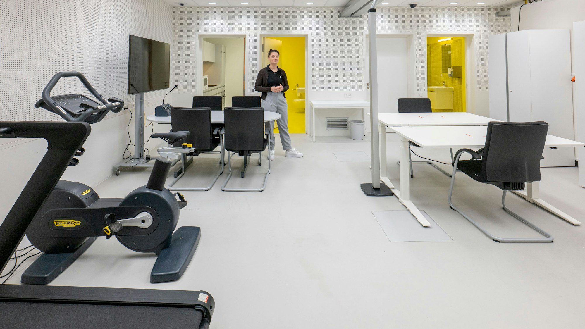 Der Aufenthaltsraum der SOLIS-Studie: Es gibt ein Laufband, ein Fahrradergometer, zwei Tische und einen Fernseher. Im Hintergrund sieht man den Eingang zu zwei Bädern und der Küche.