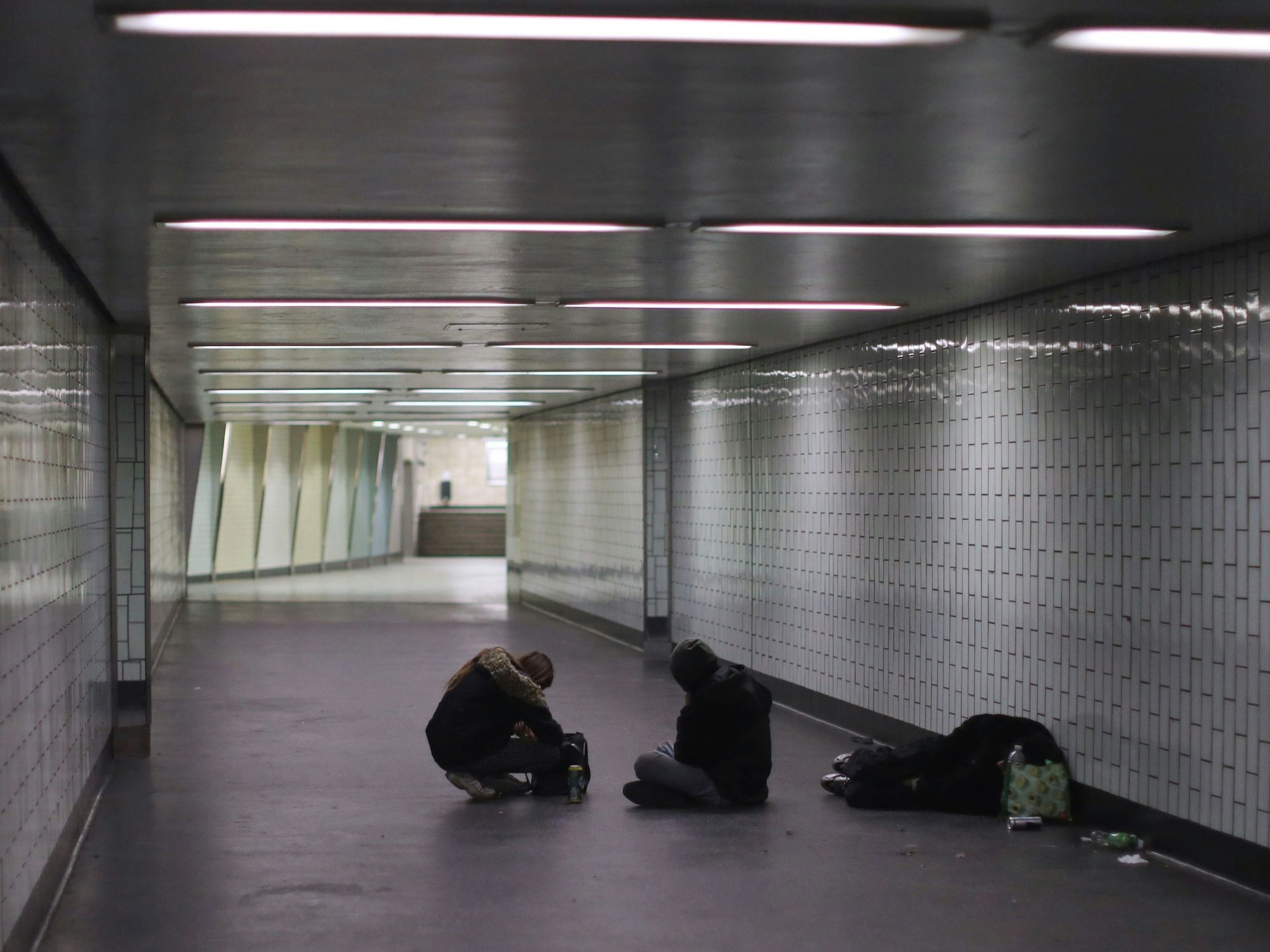 Eine Person liegt in einem Gang einer Haltestelle auf dem Boden, zwei weitere sitzen neben ihm.