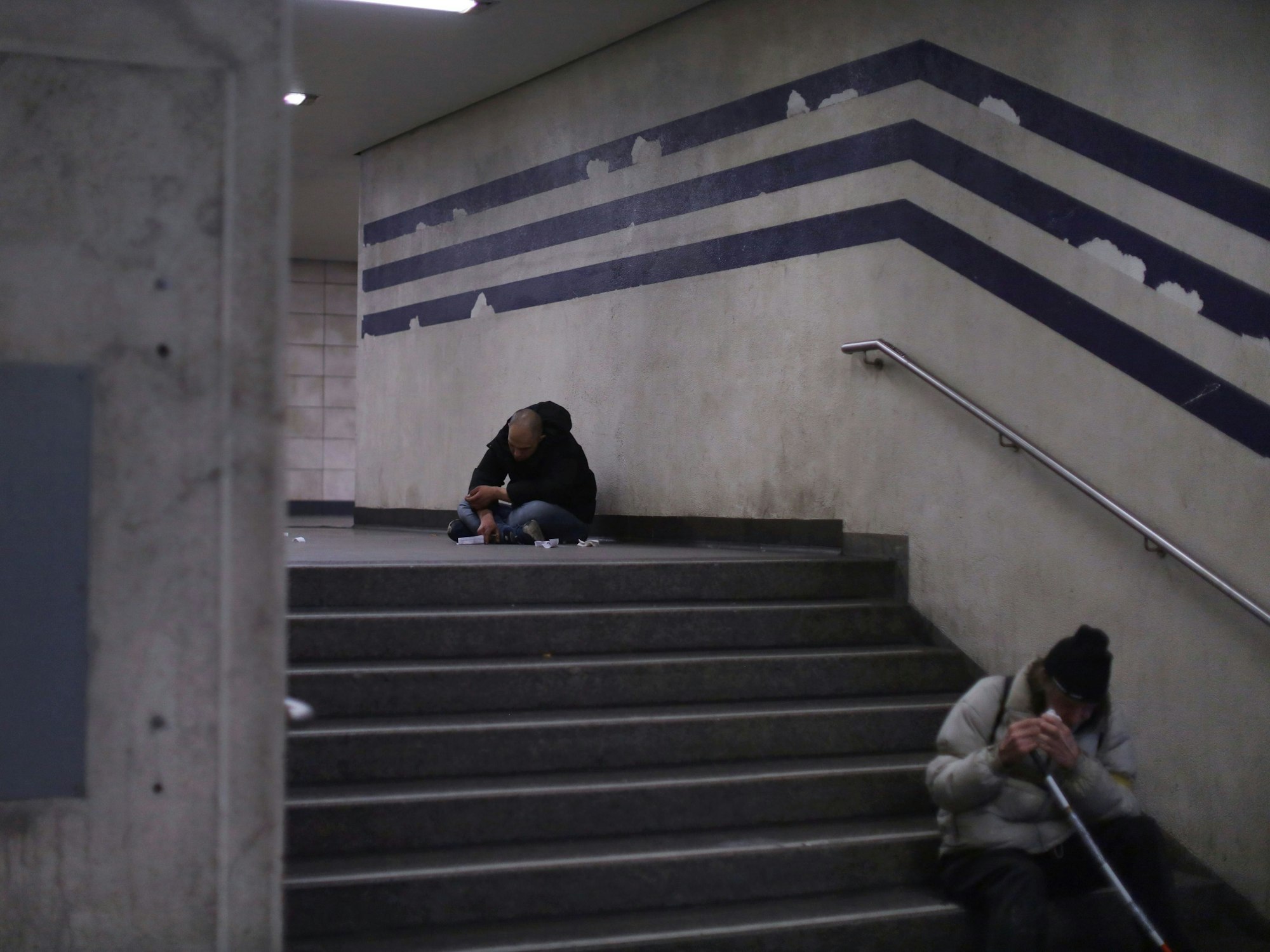 Zwei Personen sitzen zusammengesunken auf einem Treppenabsatz beziehungsweise einer Treppenstufe.