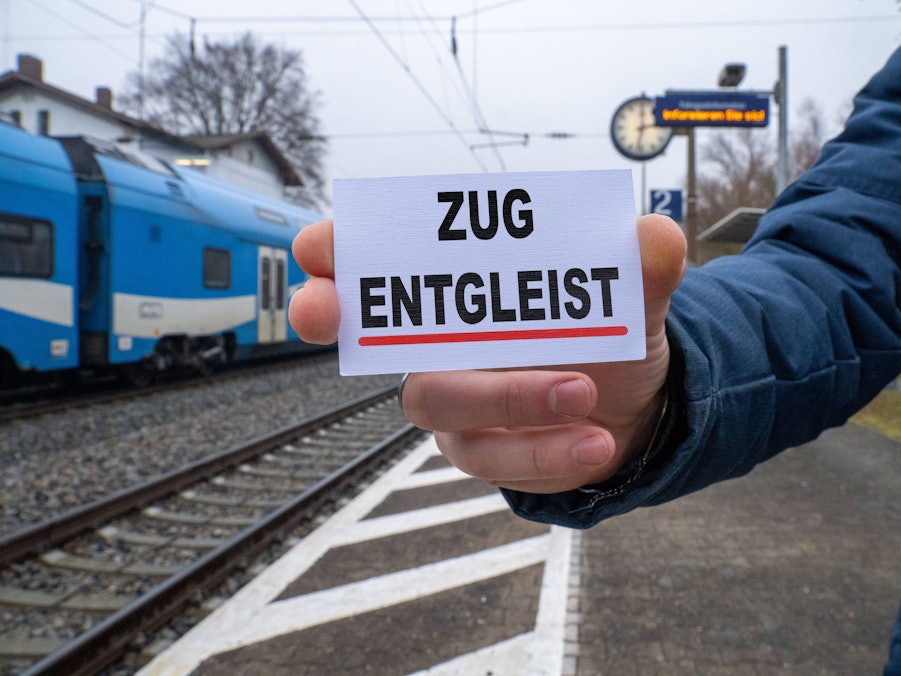 Person mit Schild „Zug entgleist“ an Bahnhof