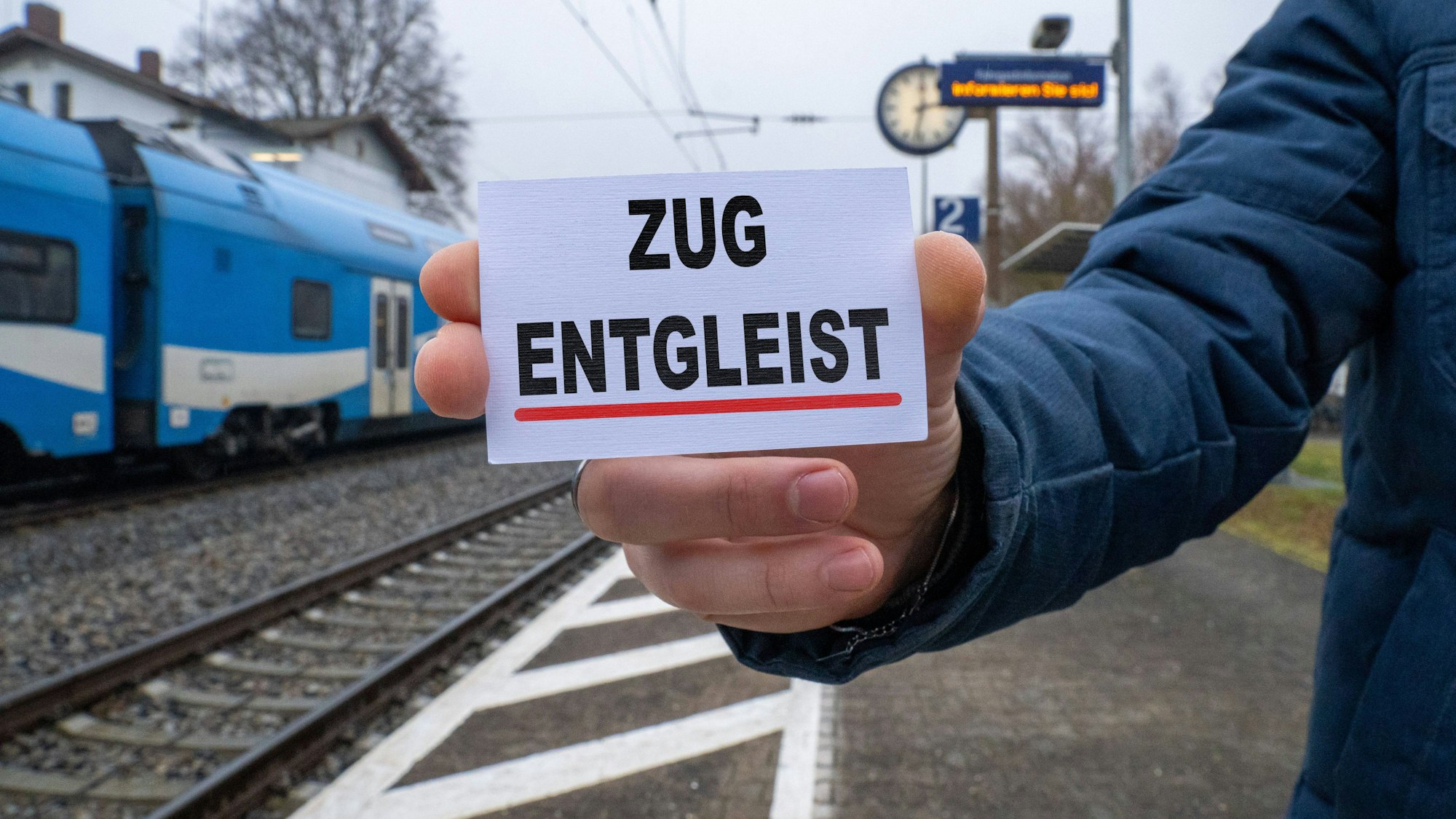 Person mit Schild „Zug entgleist“ an Bahnhof