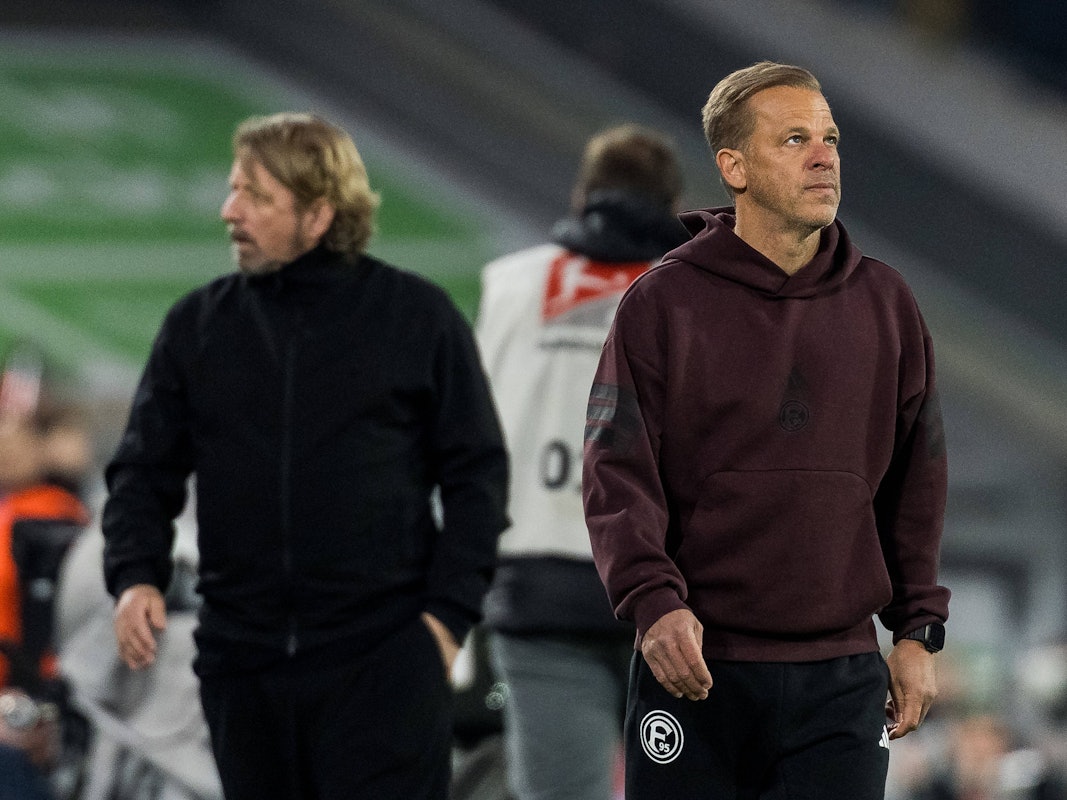 Markus Anfang schaut nachdenklich in die Luft, im Hintergrund geht Sven Mislintat.