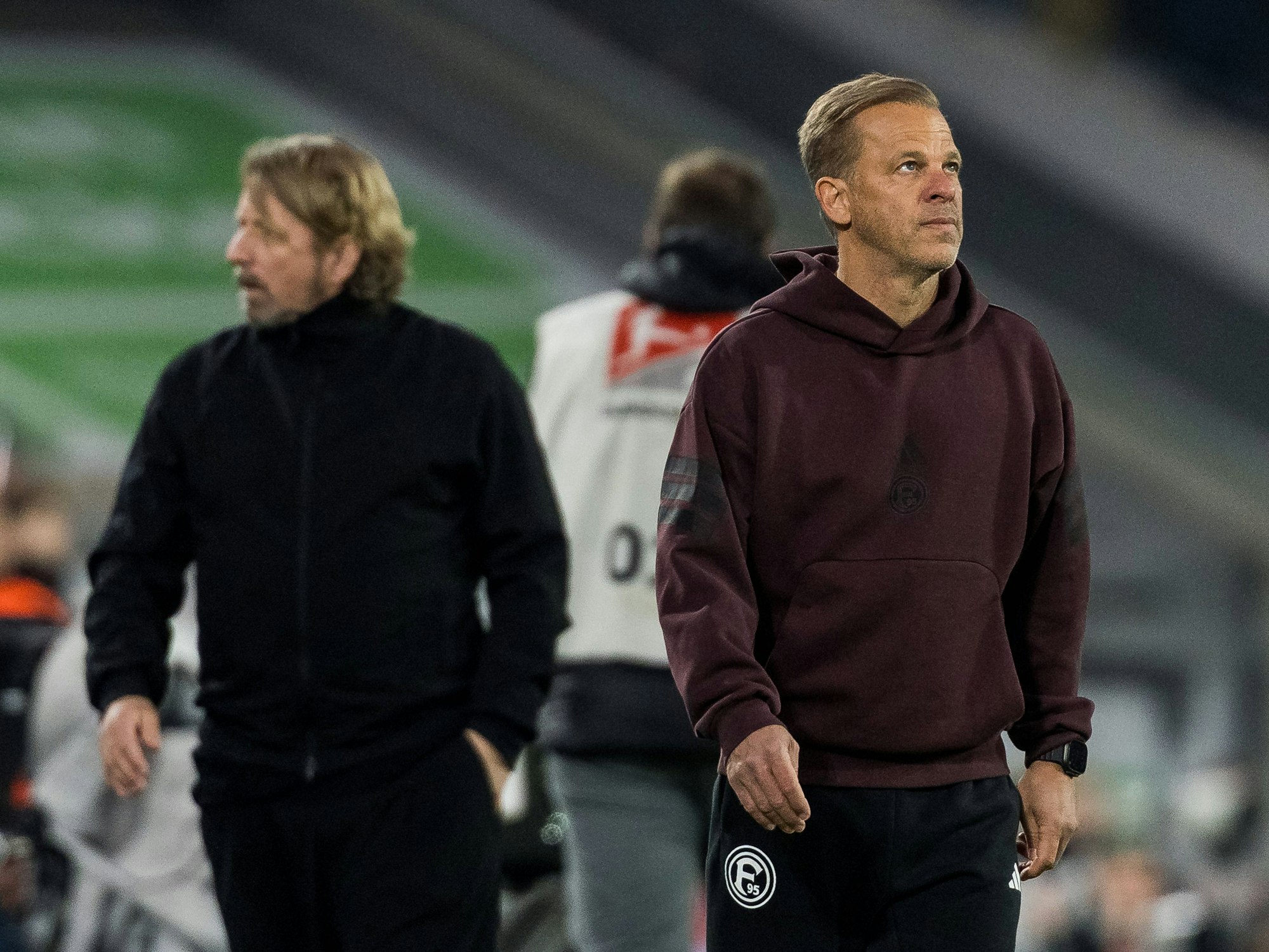 Markus Anfang schaut nachdenklich in die Luft, im Hintergrund geht Sven Mislintat.