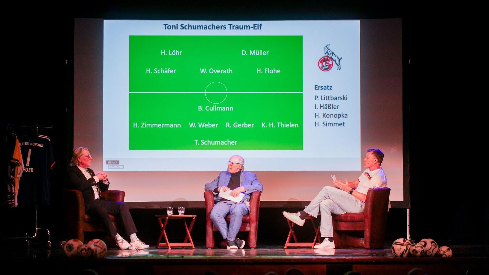 Toni Schumacher (l.) sitzt neben Reiner Calmund und Thomas Helmer (r.) auf der Bühne.