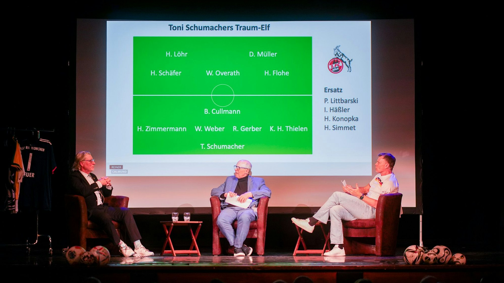 Toni Schumacher (l.) sitzt neben Reiner Calmund und Thomas Helmer (r.) auf der Bühne.