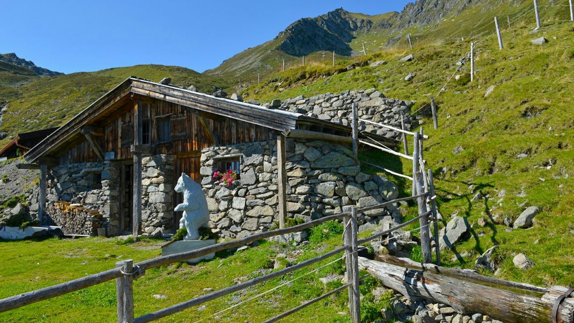 Eine kleine, aus Bruchsteinen gefertigte Almhütte schmiegt sich in die grüne Rasenflanke eines Berges.