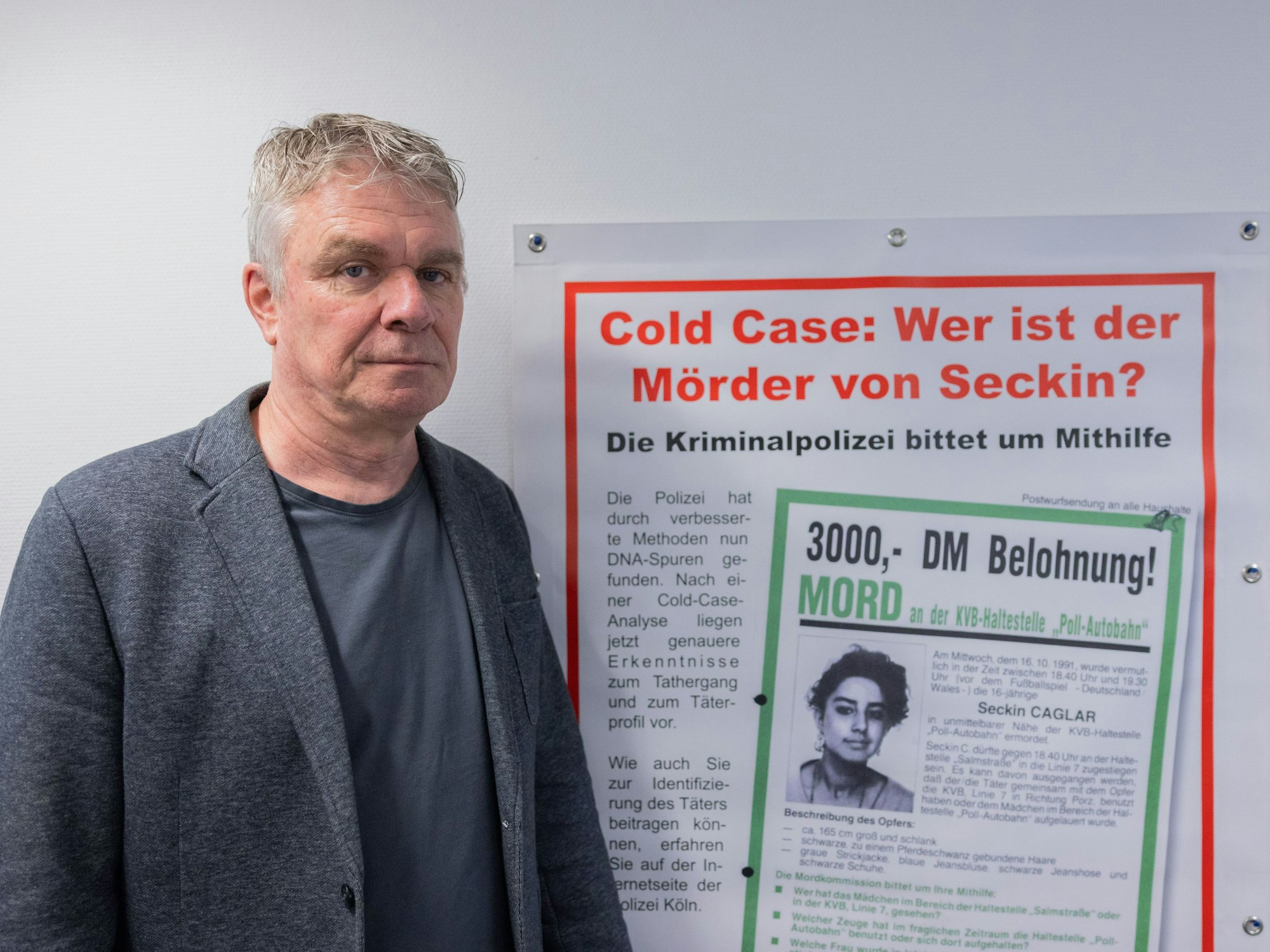 Markus Weber steht neben einem Fahndungsplakat von einem ungelösten Fall mit der Überschrift: Cold Case: Wer ist der Mörder von Seckin?