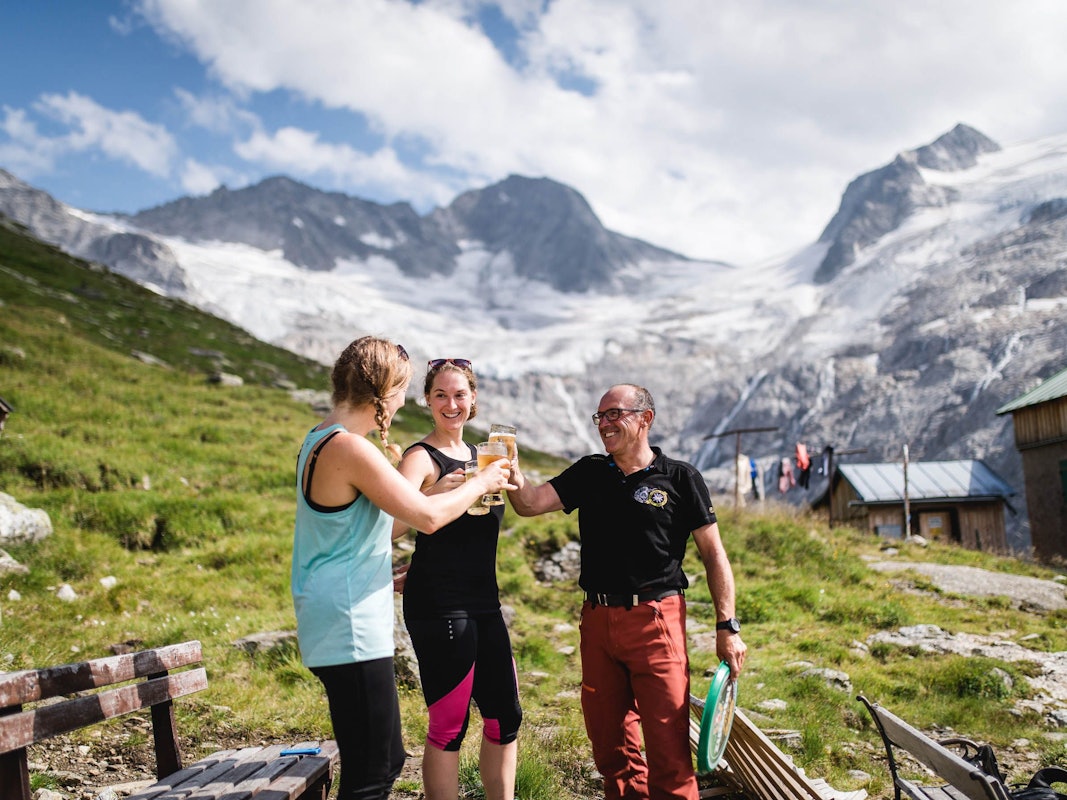 Zwei Wanderinnen stoßen mit einem Hüttenwirt auf der Terrasse einer Hütte mit Blick in die Berge an.