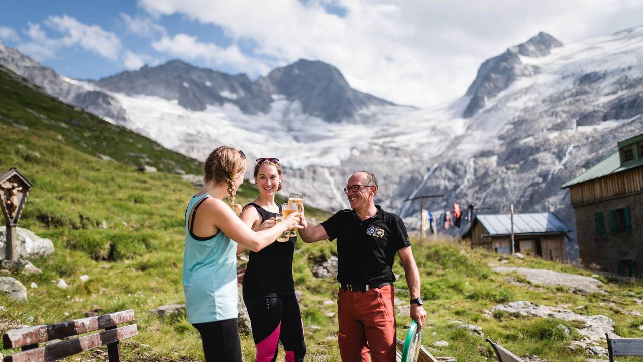 Zwei Wanderinnen stoßen mit einem Hüttenwirt auf der Terrasse einer Hütte mit Blick in die Berge an.