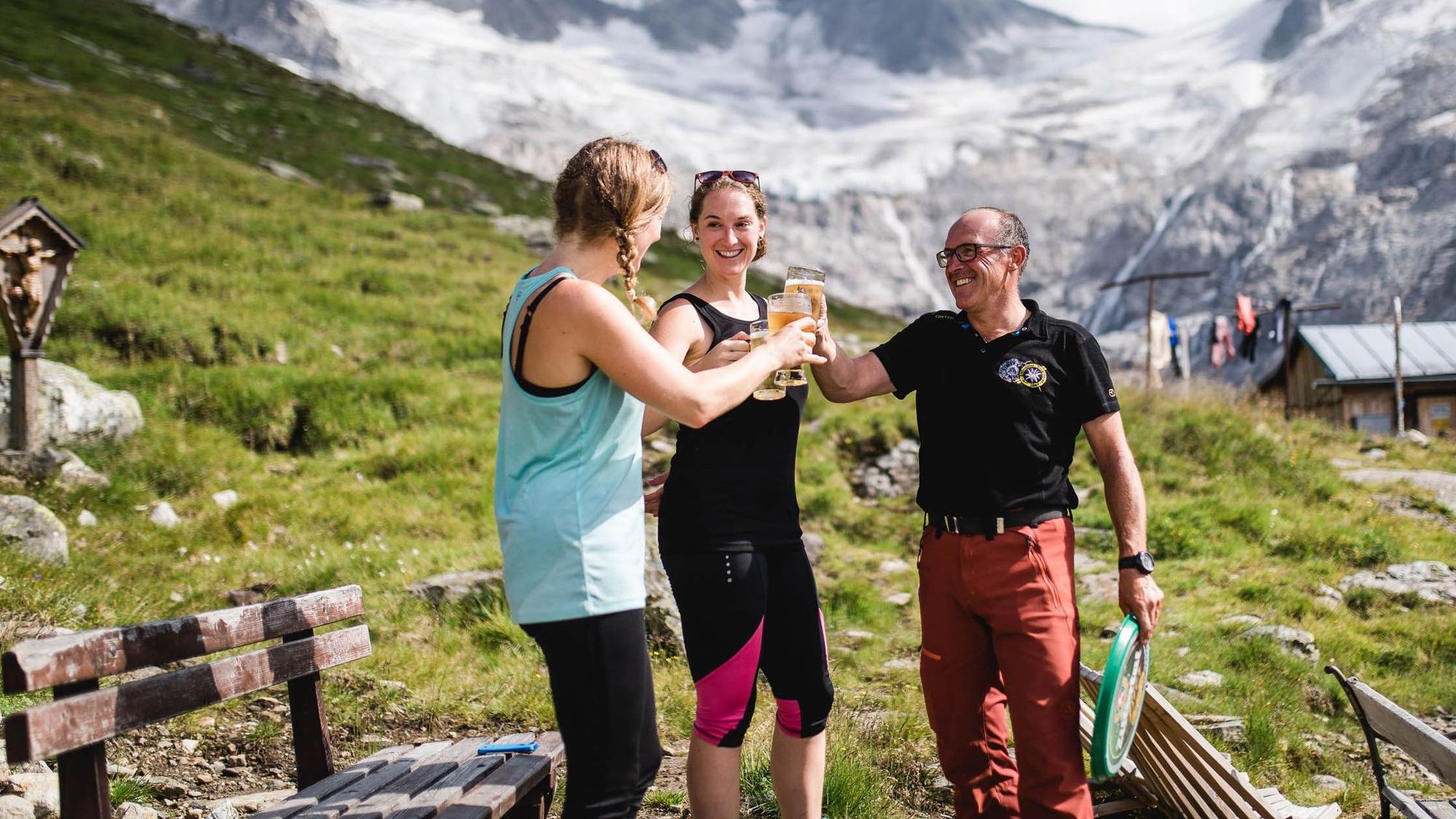 Zwei Wanderinnen stoßen mit einem Hüttenwirt auf der Terrasse einer Hütte mit Blick in die Berge an.