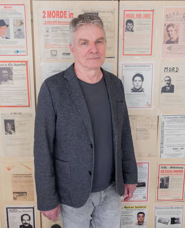 Markus Weber, Leiter Ermittlungsgruppe Cold Cases der Kölner Polizei, steht in einem Büro im Polizeipräsidium vor Büroschränken, auf denen Fahndungsplakate mit ungelösten Fällen hängen.