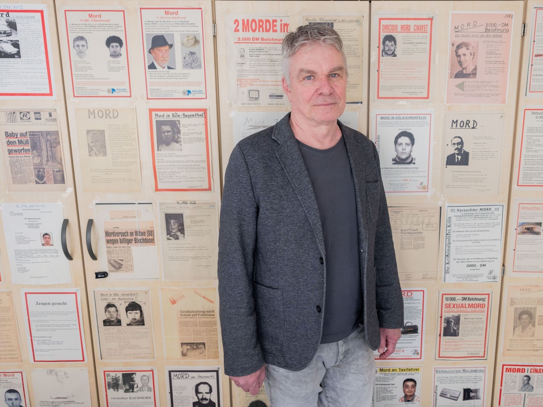 Markus Weber, Leiter Ermittlungsgruppe Cold Cases der Kölner Polizei, steht in einem Büro im Polizeipräsidium vor Büroschränken, auf denen Fahndungsplakate mit ungelösten Fällen hängen.