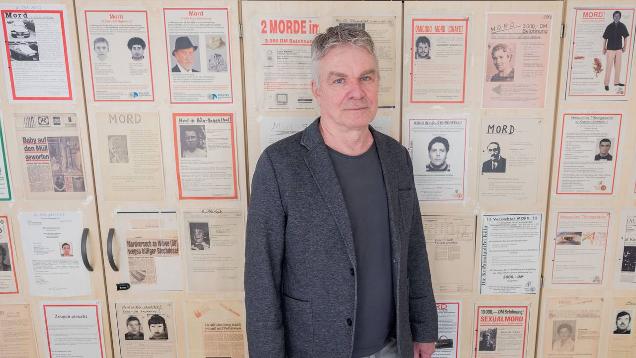 Markus Weber, Leiter Ermittlungsgruppe Cold Cases der Kölner Polizei, steht in einem Büro im Polizeipräsidium vor Büroschränken, auf denen Fahndungsplakate mit ungelösten Fällen hängen.