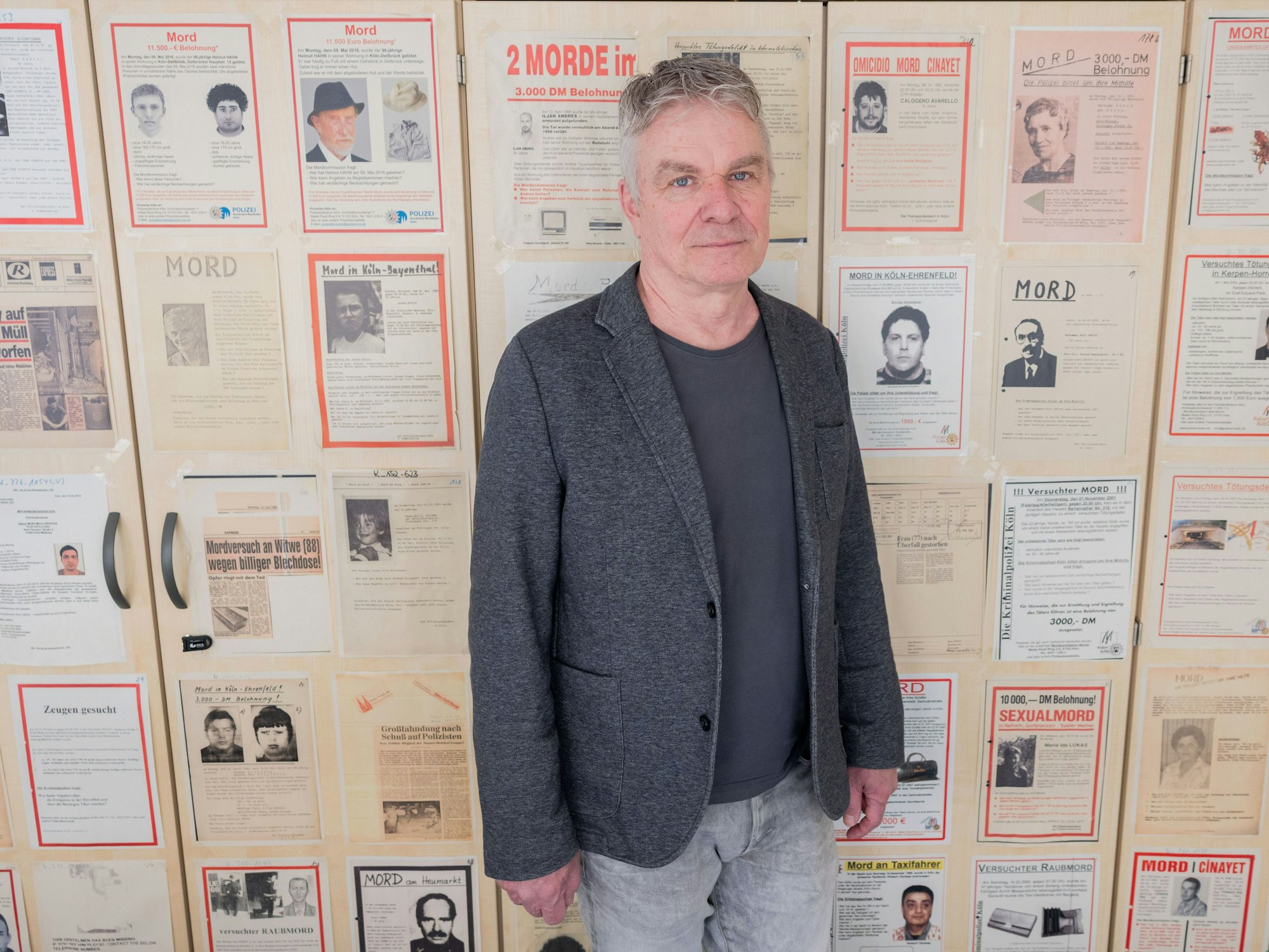 Markus Weber, Leiter Ermittlungsgruppe Cold Cases der Kölner Polizei, steht in einem Büro im Polizeipräsidium vor Büroschränken, auf denen Fahndungsplakate mit ungelösten Fällen hängen.