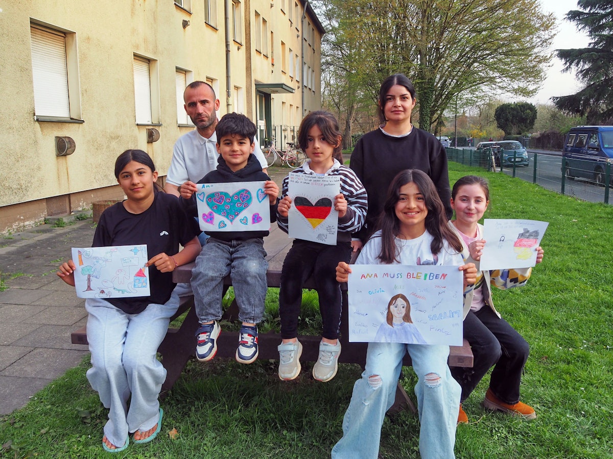 Ana und ihre Familie sitzen vor einem Mehrfamilienhaus und halten selbstgemalte Plakate in den Händen, unter anderem ein Herz in den Deutschland-Farben.