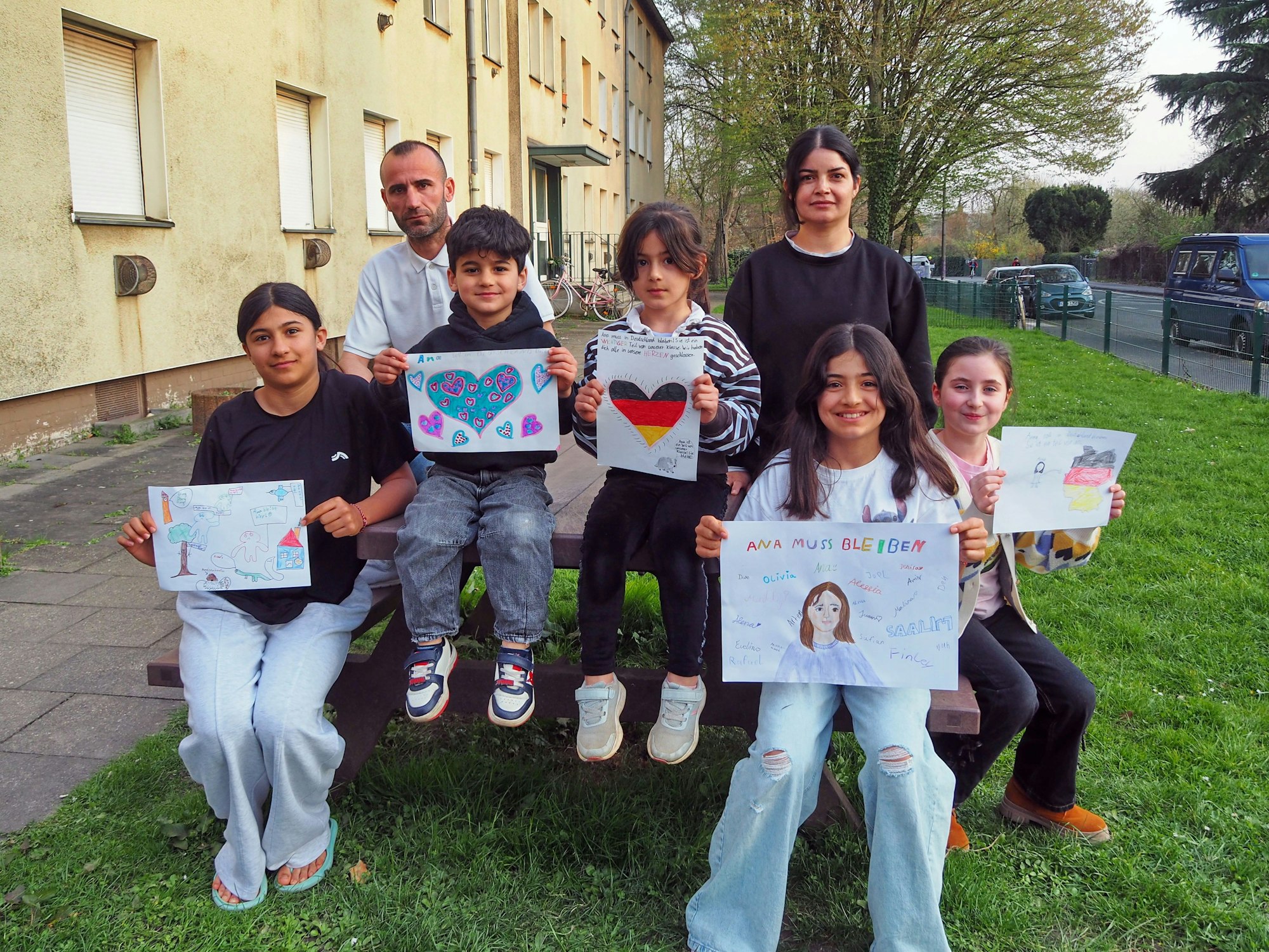 Ana und ihre Familie sitzen vor einem Mehrfamilienhaus und halten selbstgemalte Plakate in den Händen, unter anderem ein Herz in den Deutschland-Farben.