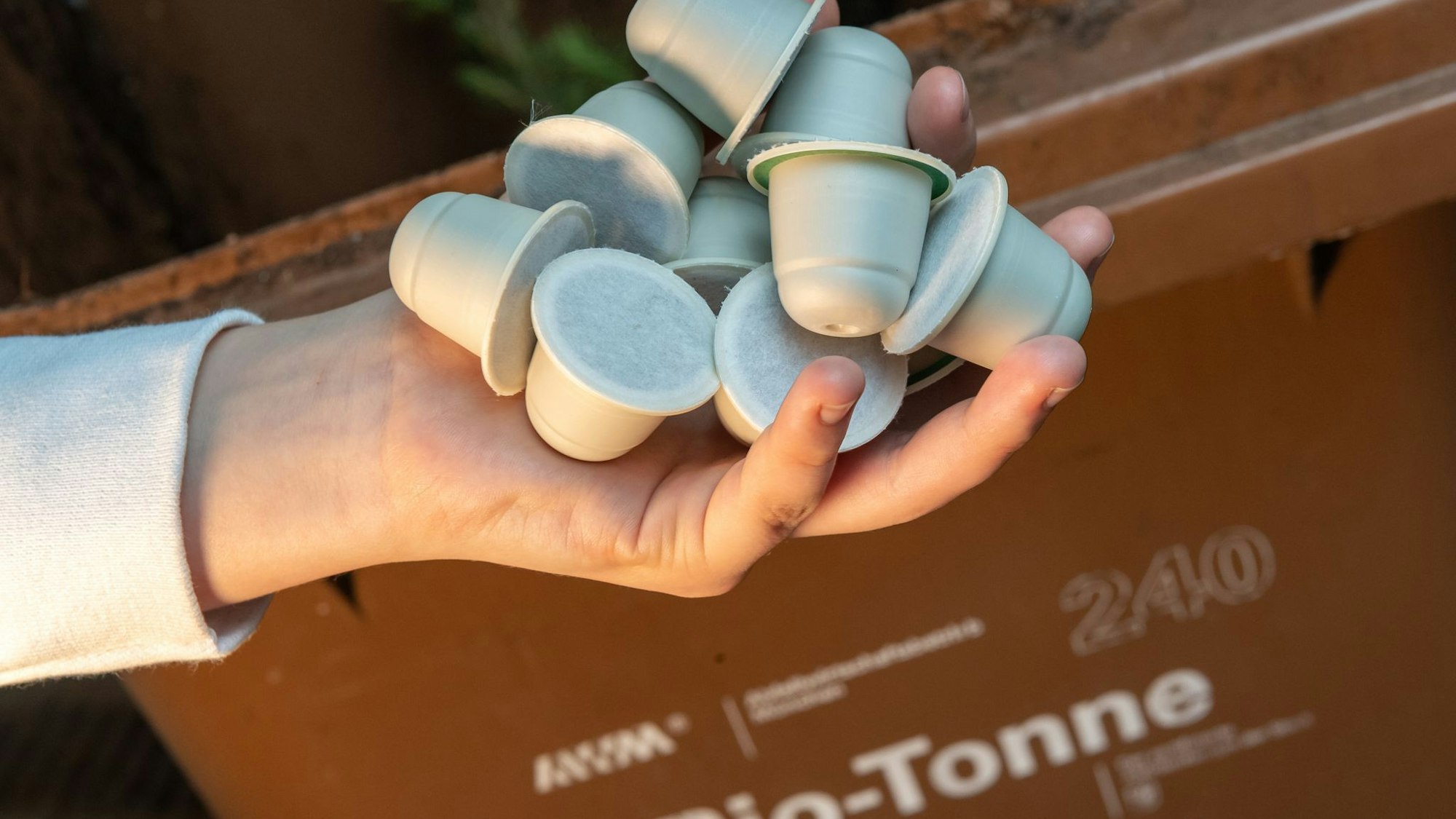 Frau hält kompostierbare Kaffeekapseln vor einer Bio-Mülltonne
