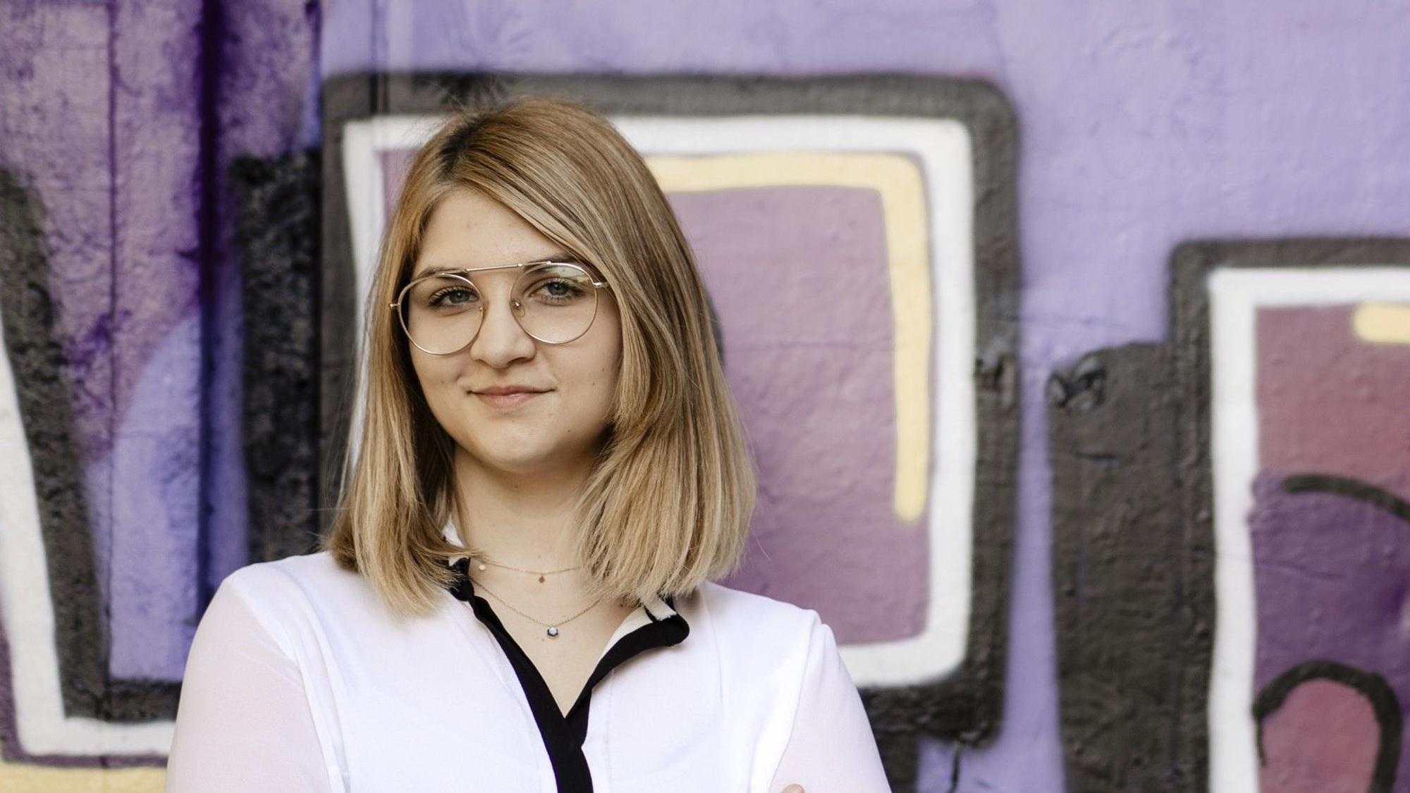 Eine junge Frau mit Brille und langen Haaren steht vor einer Graffitiwand und blickt in die Kamera