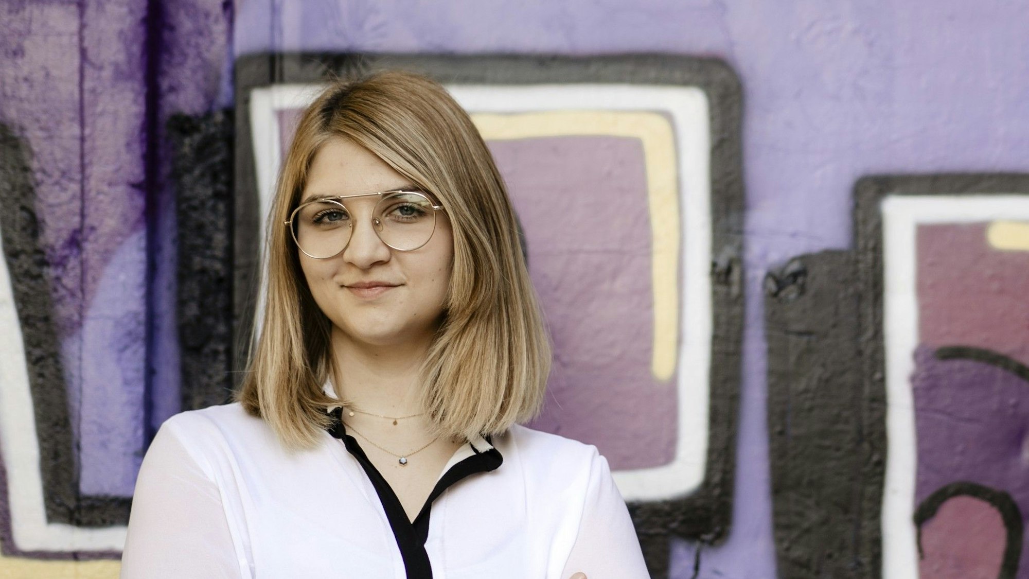 Eine junge Frau mit Brille und langen Haaren steht vor einer Graffitiwand und blickt in die Kamera