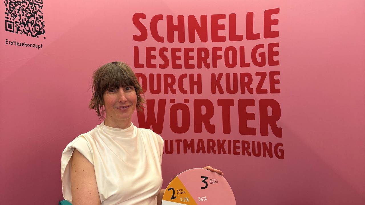 Eine Frau mit hochgesteckten Haaren in einem engen eierschalfarbenen Kleid hält eine Pappe mit einem Tortendiaframm vor einem roten Hintergrund in die Höhe.