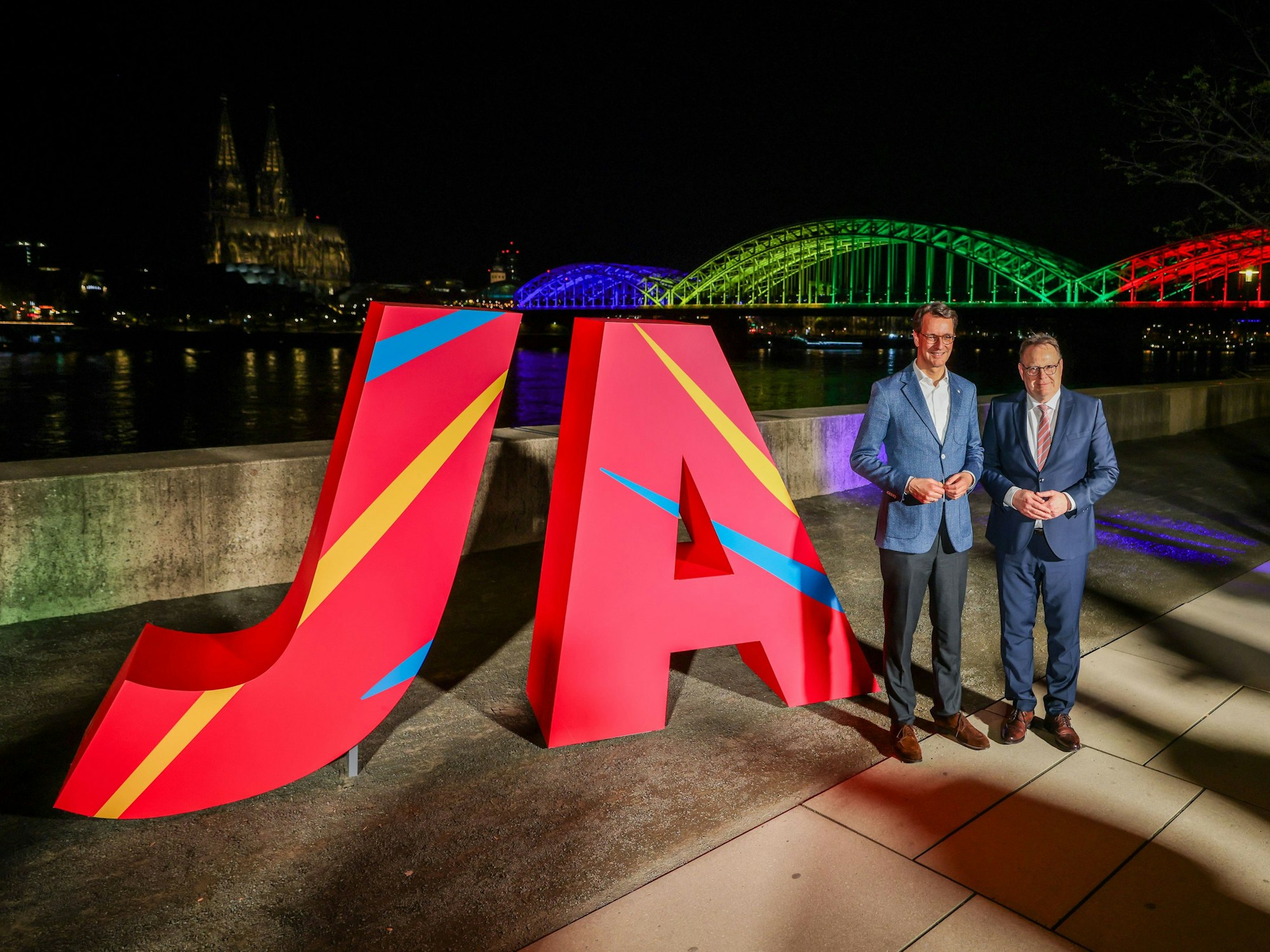 Sie feiern schon, während in Deutz noch gezählt wird: NRW-Ministerpräsident Hendrik Wüst und der Kölner Oberbürgermeister Torsten Burmester stehen vor dem großen „Ja“ und der in den olympischen Farben angeleuchteten Hohenzollernbrücke.