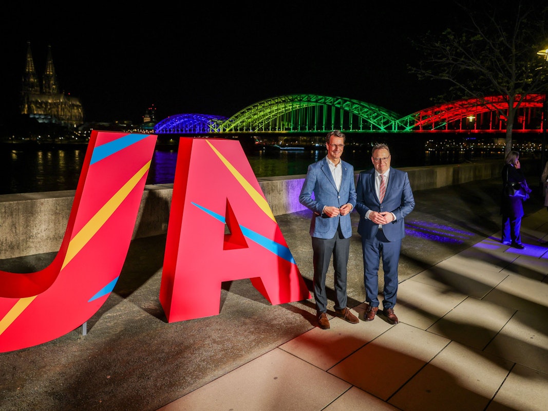 Hendrik Wüst (CDU, l.), NRW-Ministerpräsident, und Torsten Burmester (SPD), Oberbürgermeister der Stadt Köln, stehen vor dem großen „Ja“ und der in den olympischen Farben angeleuchteten Hohenzollernbrücke.