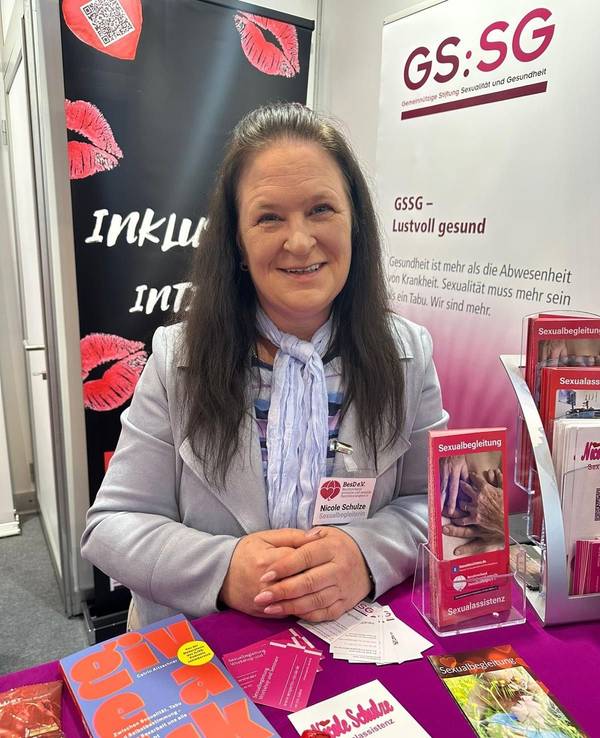 Nicole Schulze steht an einem Messestand mit Broschüren zum Thema Sexualbegleitung, hinter ihr hängt ein Banner mit Kussmündern und der Aufschrift Inklusiv Intim.