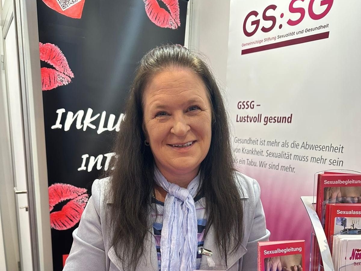 Nicole Schulze steht an einem Messestand mit Broschüren zum Thema Sexualbegleitung, hinter ihr hängt ein Banner mit Kussmündern und der Aufschrift Inklusiv Intim.