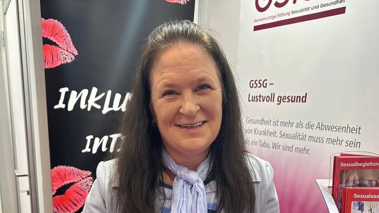 Nicole Schulze steht an einem Messestand mit Broschüren zum Thema Sexualbegleitung, hinter ihr hängt ein Banner mit Kussmündern und der Aufschrift Inklusiv Intim.