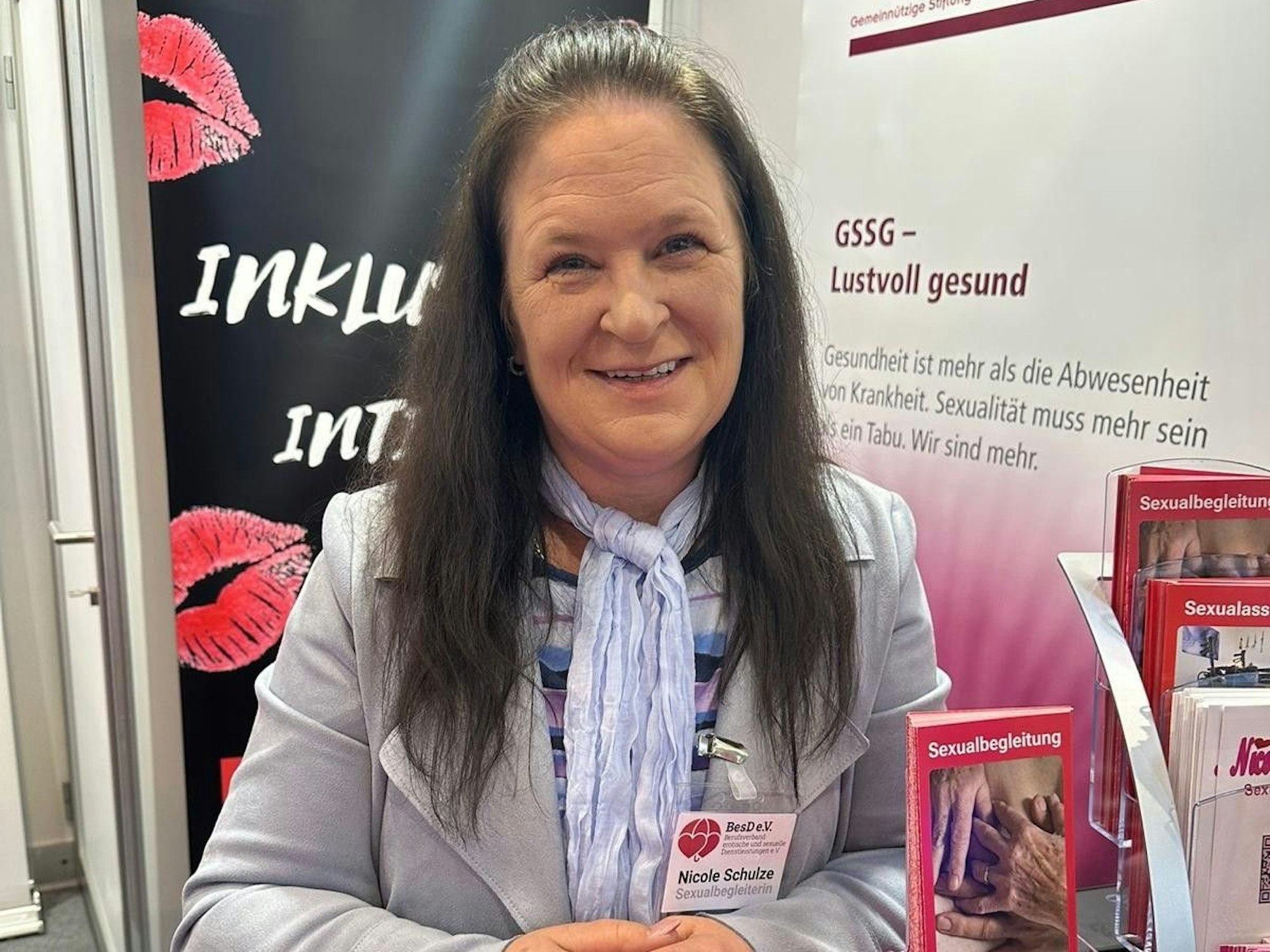 Nicole Schulze steht an einem Messestand mit Broschüren zum Thema Sexualbegleitung, hinter ihr hängt ein Banner mit Kussmündern und der Aufschrift Inklusiv Intim.