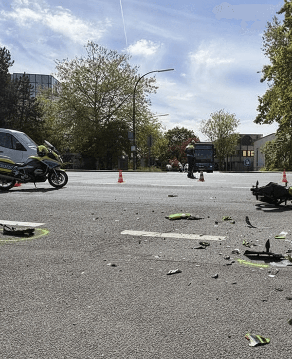 Das Foto zeigt eine Straßenszene nach einem Unfall. Man sieht ein stark beschädigtes Auto auf der rechten Seite und ein Motorrad, das auf der Straße liegt, umgeben von Trümmern.