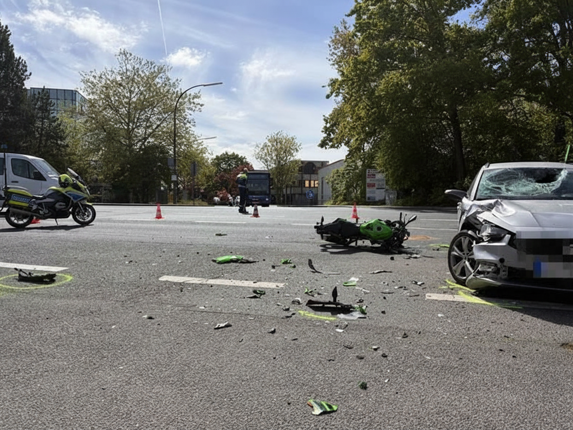 Das Foto zeigt eine Straßenszene nach einem Unfall. Man sieht ein stark beschädigtes Auto auf der rechten Seite und ein Motorrad, das auf der Straße liegt, umgeben von Trümmern.