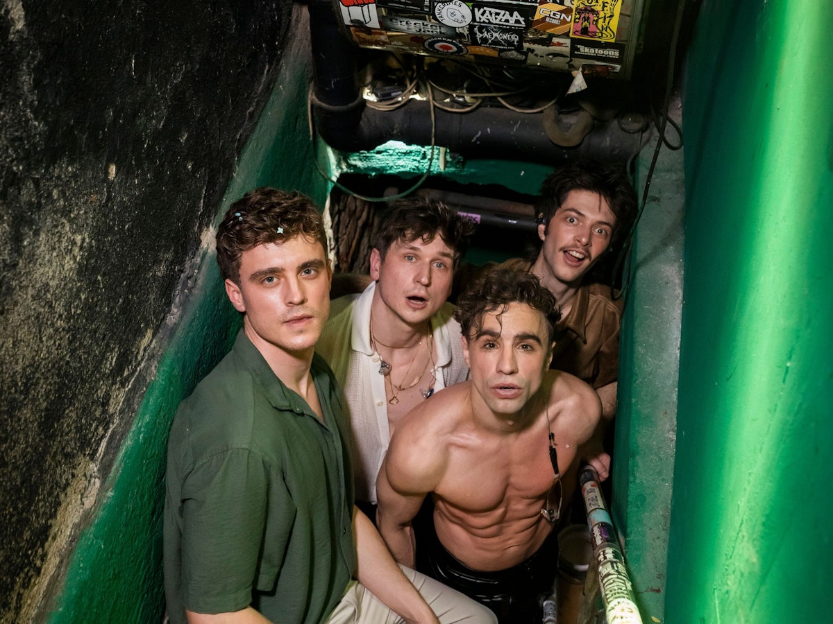 Vier junge Männer, einer mit nacktem Oberkörper, stehen in einem Treppenaufgang.