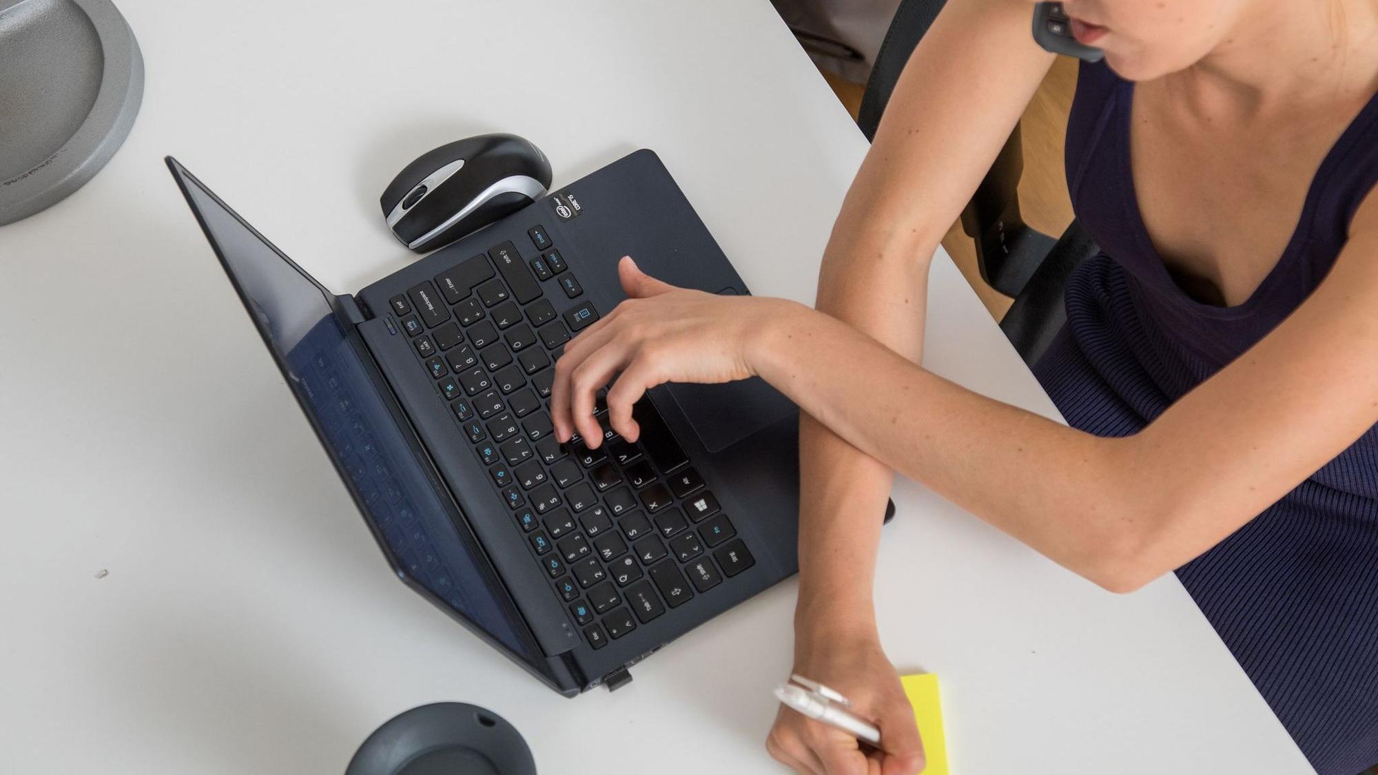 Eine Frau telefoniert, tippt auf Laptop und schreibt Notiz