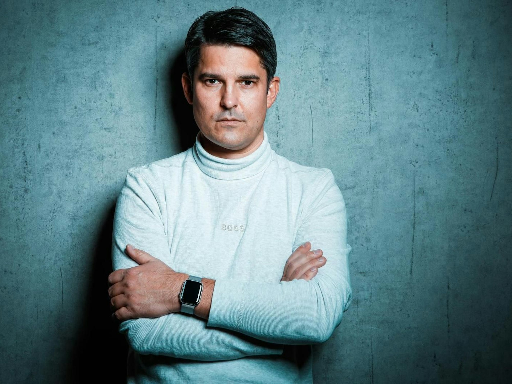 Ein Mann steht mit verschränkten Armen vor einer Wand.