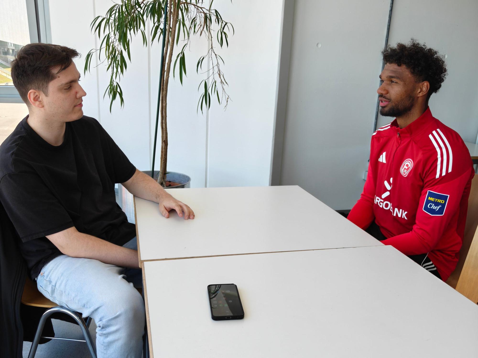 Express-Reporter Sebastian Bucco und Fortuna-Spieler Elias Egouli sitzen an einem Tisch gegenüber und sprechen miteinander.