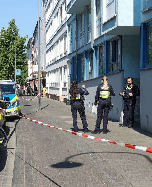 Die Polizei hat einen Bürgersteig abgesperrt, Polizeifahrzeuge parken vor dem Haus, drei Einsatzkräfte stehen vor der Haustür.