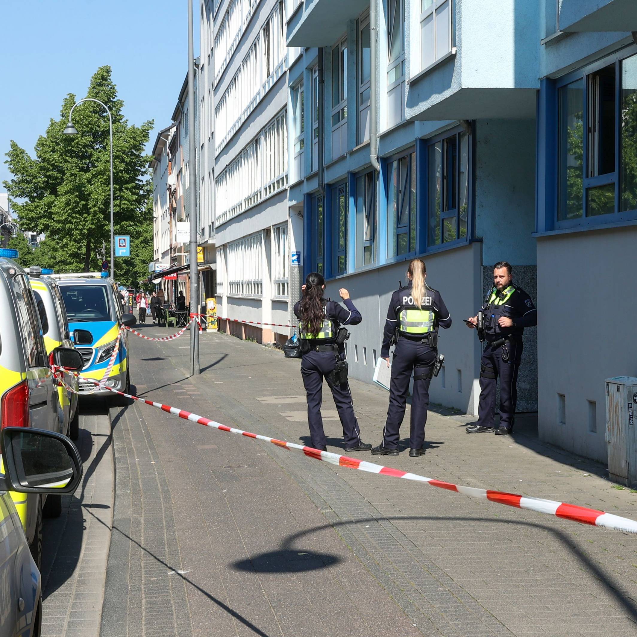 Die Polizei hat einen Bürgersteig abgesperrt, Polizeifahrzeuge parken vor dem Haus, drei Einsatzkräfte stehen vor der Haustür.