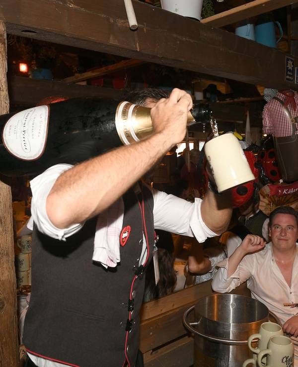 Ein Kellner hat eine Champagnerflasche auf der Schulter und schenkt einem Gast in einen Krug Champagner im Käferzelt aus.