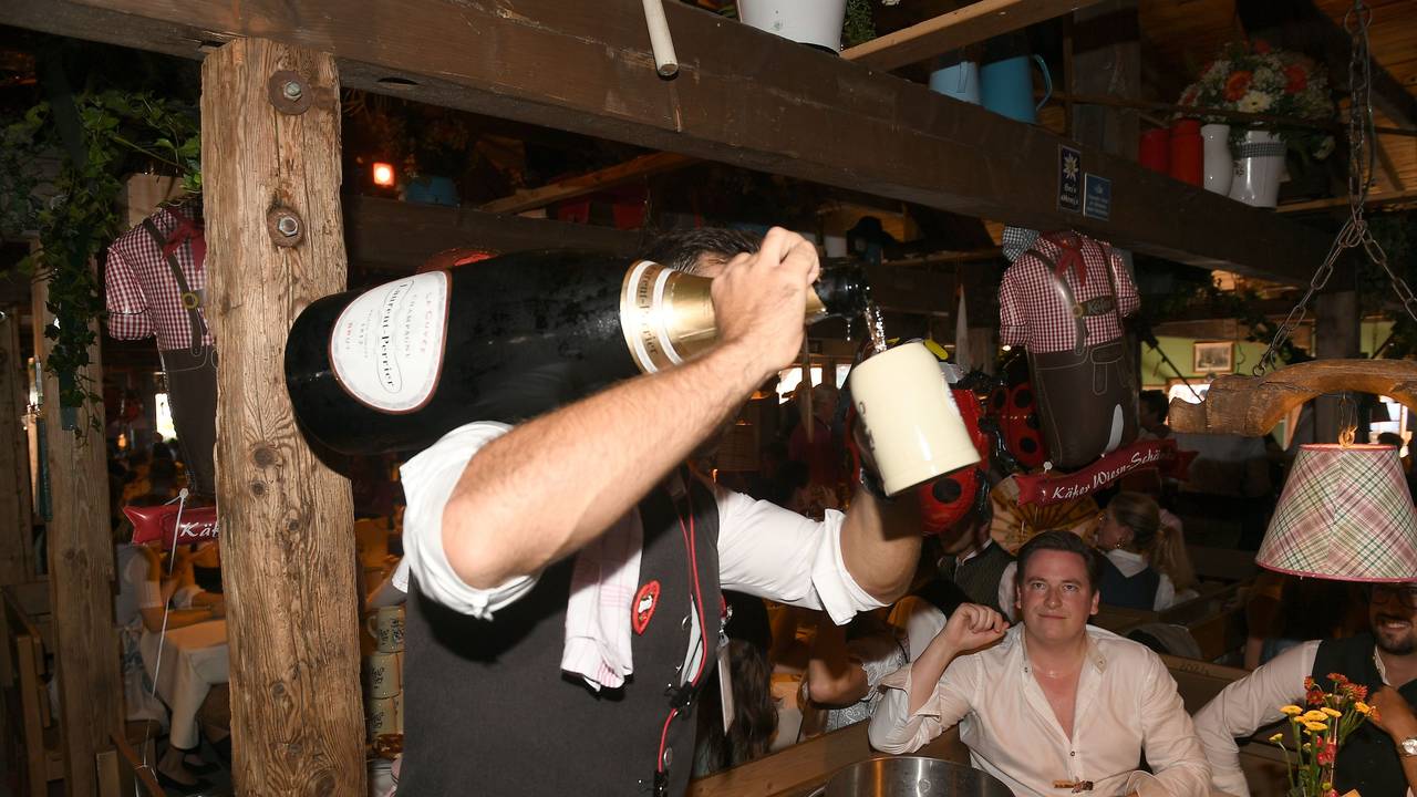 Ein Kellner hat eine Champagnerflasche auf der Schulter und schenkt einem Gast in einen Krug Champagner im Käferzelt aus.