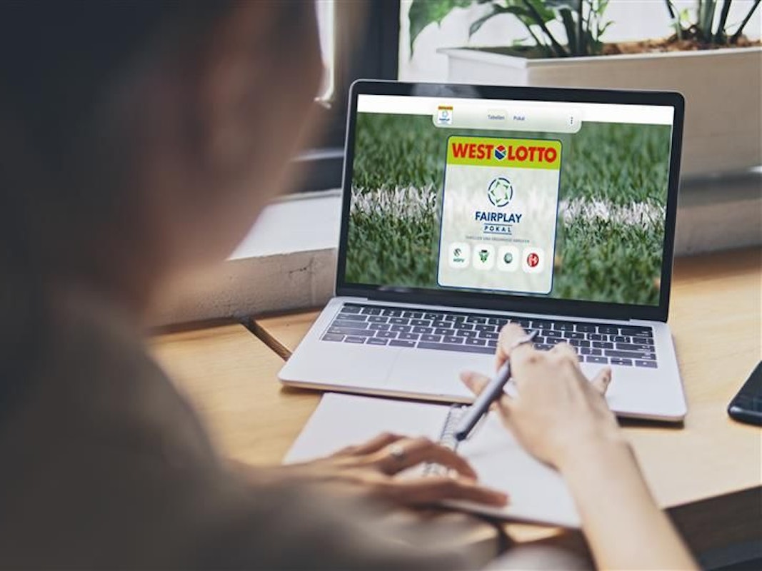 Eine Frau sitzt an einem Tisch und schaut auf ihren Laptop. Der Laptop zeigt die Internetseite des WestLotto Fairplay-Pokals.