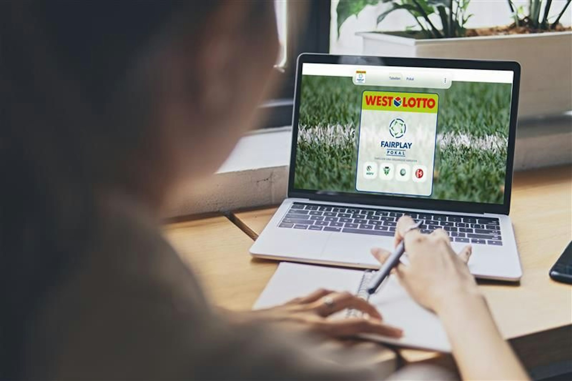 Eine Frau sitzt an einem Tisch und schaut auf ihren Laptop. Der Laptop zeigt die Internetseite des WestLotto Fairplay-Pokals.