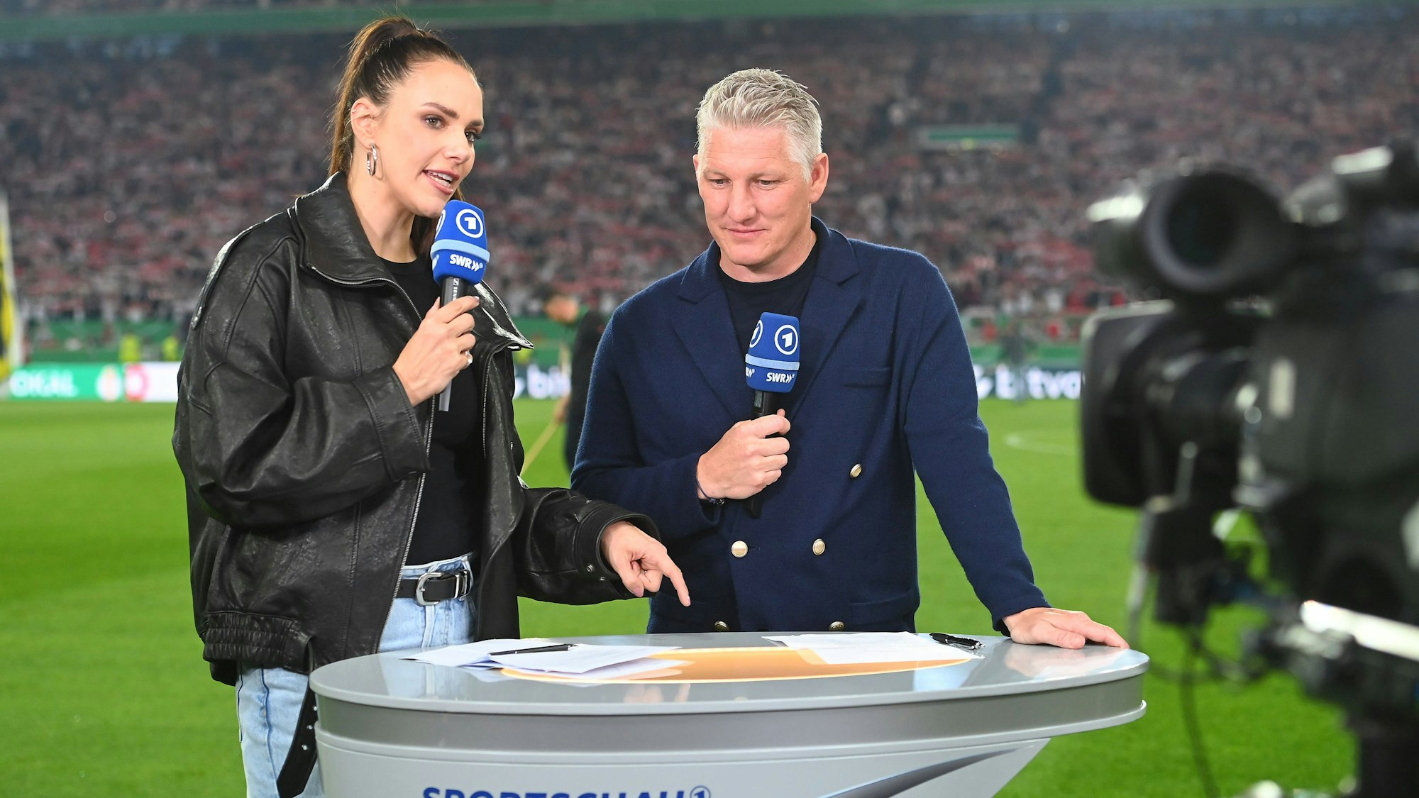 ARD-Moderatorin Esther Sedlaczek und Bastian Schweinsteiger halten bei einer TV-Übertragung Mikrofone in der Hand.