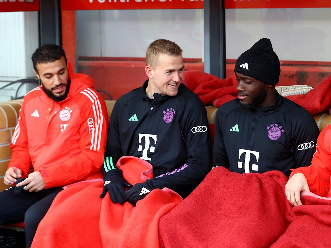 Noussair Mazraoui (l.), Matthijs de Ligt (M.) und Dayot Upamecano sitzen auf der Ersatzbank bei einem Spiel des FC Bayern.